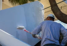 Homem com camiseta azul e boné branco está em cima de uma escada, segurando um rolo de pintura com cabo vermelho, enquanto pinta uma parede de azul claro.