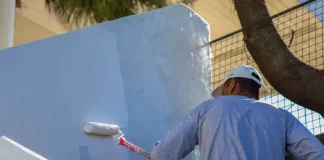 Homem com camiseta azul e boné branco está em cima de uma escada, segurando um rolo de pintura com cabo vermelho, enquanto pinta uma parede de azul claro.