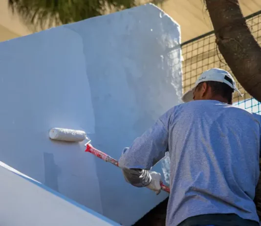 Estado reduz custos e qualifica internos com reforma de hospitais feita por apenados Homem com camiseta azul e boné branco está em cima de uma escada, segurando um rolo de pintura com cabo vermelho, enquanto pinta uma parede de azul claro.