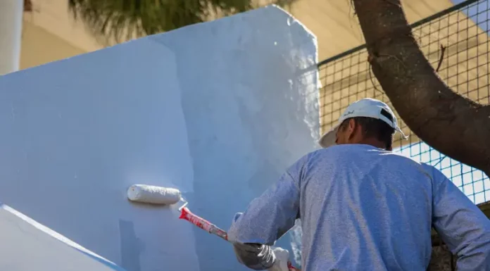 Homem com camiseta azul e boné branco está em cima de uma escada, segurando um rolo de pintura com cabo vermelho, enquanto pinta uma parede de azul claro.