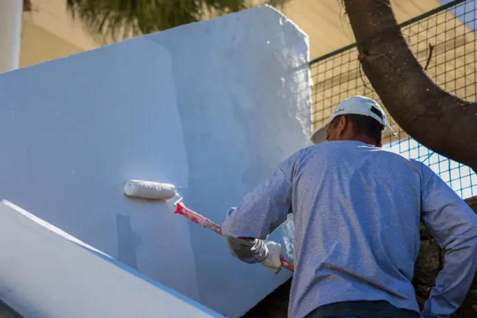 Homem com camiseta azul e boné branco está em cima de uma escada, segurando um rolo de pintura com cabo vermelho, enquanto pinta uma parede de azul claro.