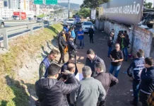 Na foto, várias pessoas estão vistoriando uma área vulnerável em um beco estreito, localizado ao lado da Via Expressa, em São José. O grupo caminha e observa o local, que tem vegetação rasteira,e algumas estruturas precárias. O ambiente transmite sensação de abandono e vulnerabilidade.