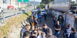 Na foto, várias pessoas estão vistoriando uma área vulnerável em um beco estreito, localizado ao lado da Via Expressa, em São José. O grupo caminha e observa o local, que tem vegetação rasteira,e algumas estruturas precárias. O ambiente transmite sensação de abandono e vulnerabilidade.