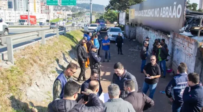 Na foto, várias pessoas estão vistoriando uma área vulnerável em um beco estreito, localizado ao lado da Via Expressa, em São José. O grupo caminha e observa o local, que tem vegetação rasteira,e algumas estruturas precárias. O ambiente transmite sensação de abandono e vulnerabilidade.