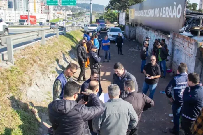 Na foto, várias pessoas estão vistoriando uma área vulnerável em um beco estreito, localizado ao lado da Via Expressa, em São José. O grupo caminha e observa o local, que tem vegetação rasteira,e algumas estruturas precárias. O ambiente transmite sensação de abandono e vulnerabilidade.