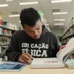 Aluno beneficiado pelo programa Universidade Gratuita, vestindo moletom marrom com a inscrição "Educação Física", consulta um livro enquanto estuda em uma biblioteca.