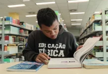Aluno beneficiado pelo programa Universidade Gratuita, vestindo moletom marrom com a inscrição "Educação Física", consulta um livro enquanto estuda em uma biblioteca.