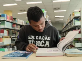 Aluno beneficiado pelo programa Universidade Gratuita, vestindo moletom marrom com a inscrição "Educação Física", consulta um livro enquanto estuda em uma biblioteca.