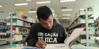 Aluno beneficiado pelo programa Universidade Gratuita, vestindo moletom marrom com a inscrição "Educação Física", consulta um livro enquanto estuda em uma biblioteca.
