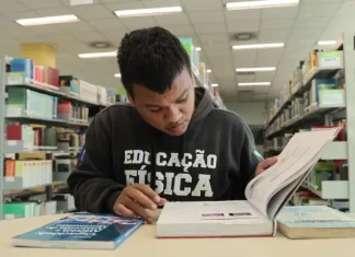 Aluno beneficiado pelo programa Universidade Gratuita, vestindo moletom marrom com a inscrição "Educação Física", consulta um livro enquanto estuda em uma biblioteca.