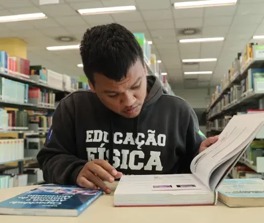 Formados pelo Universidade Gratuita começam a cumprir contrapartida com trabalho social Aluno beneficiado pelo programa Universidade Gratuita, vestindo moletom marrom com a inscrição "Educação Física", consulta um livro enquanto estuda em uma biblioteca.