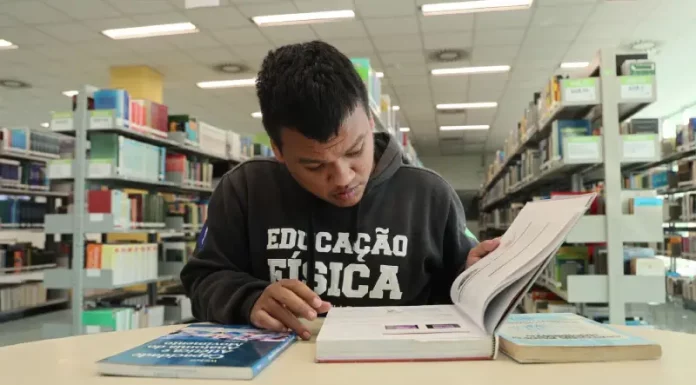 Aluno beneficiado pelo programa Universidade Gratuita, vestindo moletom marrom com a inscrição "Educação Física", consulta um livro enquanto estuda em uma biblioteca.