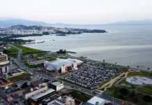 Vista aérea da cidade de São José, Santa Catarina. A imagem mostra a avenida Beira-Mar com vários carros em movimento. Ao redor da avenida, há prédios residenciais e comerciais de diferentes alturas. Em destaque, o Centro Multiuso de São José aparece próximo à orla. Ao fundo, a paisagem se estende até o mar, que aparece calmo sob um céu nublado, com nuvens cinzentas cobrindo o dia.