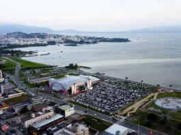 São José busca parceria para projeto de Smart City Vista aérea da cidade de São José, Santa Catarina. A imagem mostra a avenida Beira-Mar com vários carros em movimento. Ao redor da avenida, há prédios residenciais e comerciais de diferentes alturas. Em destaque, o Centro Multiuso de São José aparece próximo à orla. Ao fundo, a paisagem se estende até o mar, que aparece calmo sob um céu nublado, com nuvens cinzentas cobrindo o dia.