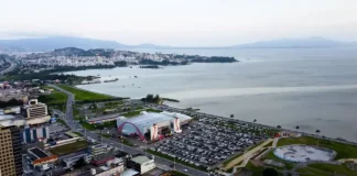 Vista aérea da cidade de São José, Santa Catarina. A imagem mostra a avenida Beira-Mar com vários carros em movimento. Ao redor da avenida, há prédios residenciais e comerciais de diferentes alturas. Em destaque, o Centro Multiuso de São José aparece próximo à orla. Ao fundo, a paisagem se estende até o mar, que aparece calmo sob um céu nublado, com nuvens cinzentas cobrindo o dia.