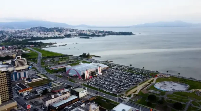 Vista aérea da cidade de São José, Santa Catarina. A imagem mostra a avenida Beira-Mar com vários carros em movimento. Ao redor da avenida, há prédios residenciais e comerciais de diferentes alturas. Em destaque, o Centro Multiuso de São José aparece próximo à orla. Ao fundo, a paisagem se estende até o mar, que aparece calmo sob um céu nublado, com nuvens cinzentas cobrindo o dia.