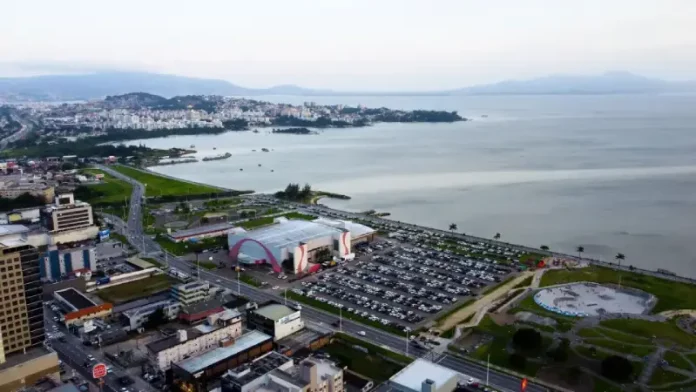 Vista aérea da cidade de São José, Santa Catarina. A imagem mostra a avenida Beira-Mar com vários carros em movimento. Ao redor da avenida, há prédios residenciais e comerciais de diferentes alturas. Em destaque, o Centro Multiuso de São José aparece próximo à orla. Ao fundo, a paisagem se estende até o mar, que aparece calmo sob um céu nublado, com nuvens cinzentas cobrindo o dia.