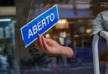 A mão de uma pessoa segura uma pequena placa azul, pendurada no lado interno do vidro de uma loja. Na placa, está escrito "ABERTO" em letras brancas e maiúsculas. A luz natural do dia ilumina o ambiente, sugerindo que a loja está pronta para receber clientes.