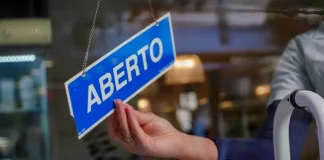 A mão de uma pessoa segura uma pequena placa azul, pendurada no lado interno do vidro de uma loja. Na placa, está escrito "ABERTO" em letras brancas e maiúsculas. A luz natural do dia ilumina o ambiente, sugerindo que a loja está pronta para receber clientes.