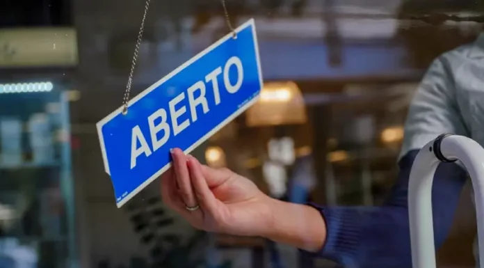 A mão de uma pessoa segura uma pequena placa azul, pendurada no lado interno do vidro de uma loja. Na placa, está escrito "ABERTO" em letras brancas e maiúsculas. A luz natural do dia ilumina o ambiente, sugerindo que a loja está pronta para receber clientes.