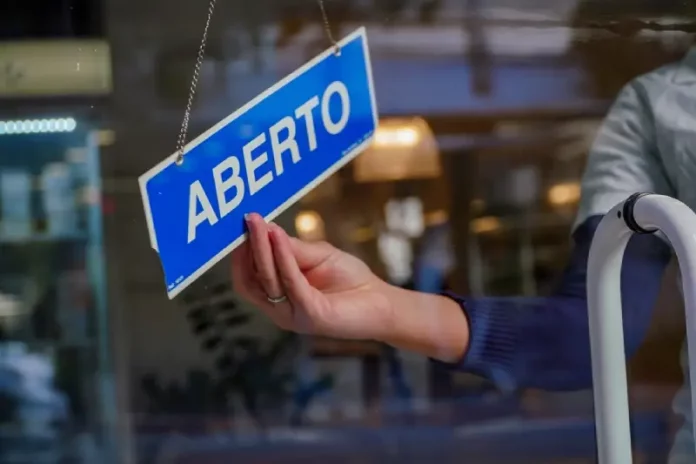 A mão de uma pessoa segura uma pequena placa azul, pendurada no lado interno do vidro de uma loja. Na placa, está escrito 