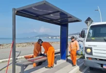 Três homens com uniformes laranja recolhem estilhaços de vidro de um abrigo de ônibus vandalizado na Beira-Mar de São José. Ao lado deles, um caminhão da prefeitura está estacionado. Ao fundo, é possível ver o mar.