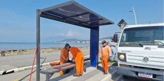 Três homens com uniformes laranja recolhem estilhaços de vidro de um abrigo de ônibus vandalizado na Beira-Mar de São José. Ao lado deles, um caminhão da prefeitura está estacionado. Ao fundo, é possível ver o mar.