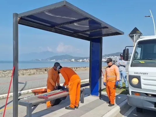 Três homens com uniformes laranja recolhem estilhaços de vidro de um abrigo de ônibus vandalizado na Beira-Mar de São José. Ao lado deles, um caminhão da prefeitura está estacionado. Ao fundo, é possível ver o mar.