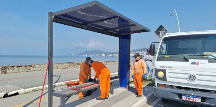Três homens com uniformes laranja recolhem estilhaços de vidro de um abrigo de ônibus vandalizado na Beira-Mar de São José. Ao lado deles, um caminhão da prefeitura está estacionado. Ao fundo, é possível ver o mar.
