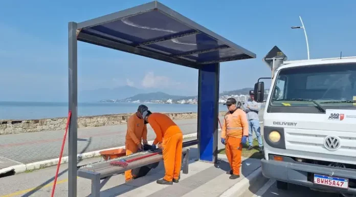 Três homens com uniformes laranja recolhem estilhaços de vidro de um abrigo de ônibus vandalizado na Beira-Mar de São José. Ao lado deles, um caminhão da prefeitura está estacionado. Ao fundo, é possível ver o mar.