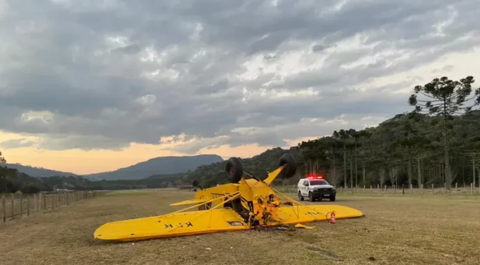 Avião de pequeno porte, na cor amarela, está virado de cabeça para baixo em um campo de vegetação baixa. Ao lado da aeronave, há um carro da polícia estacionado, com as luzes de emergência ligadas