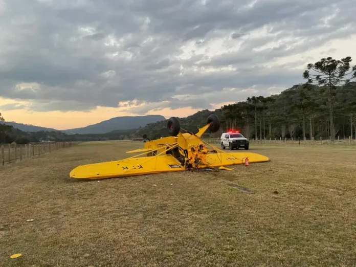 Avião de pequeno porte, na cor amarela, está virado de cabeça para baixo em um campo de vegetação baixa. Ao lado da aeronave, há um carro da polícia estacionado, com as luzes de emergência ligadas