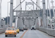 Carros antigos atravessam a Ponte Hercílio Luz, em Florianópolis, durante uma carreata. À frente, está um Karmann Ghia laranja, do governador Jorginho Mello, seguido por uma fila de veículos clássicos. A cena destaca o visual retrô dos automóveis contra a estrutura metálica da ponte e o céu parcialmente nublado ao fundo.