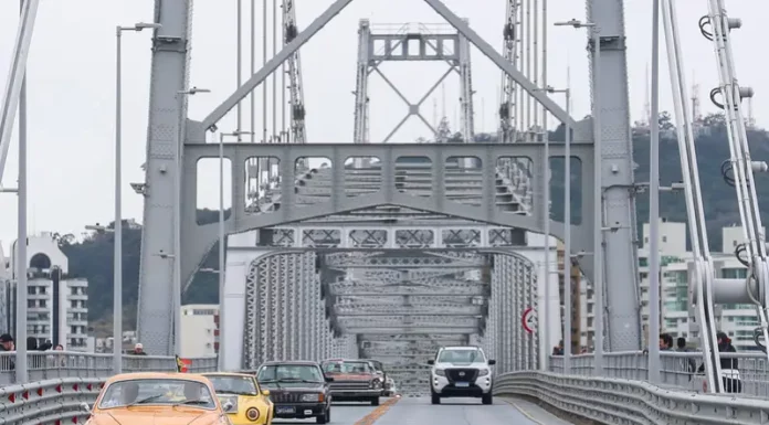Carros antigos atravessam a Ponte Hercílio Luz, em Florianópolis, durante uma carreata. À frente, está um Karmann Ghia laranja, do governador Jorginho Mello, seguido por uma fila de veículos clássicos. A cena destaca o visual retrô dos automóveis contra a estrutura metálica da ponte e o céu parcialmente nublado ao fundo.