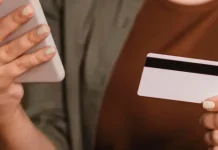 Foto mostra o tronco e as mãos de uma mulher segurando um cartão de crédito na mão esquerda e um smartphone na mão direita. A imagem simboliza o uso de tecnologia para pagamentos digitais, com foco nos objetos e nas mãos, sem mostrar o rosto da pessoa.