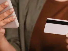 Foto mostra o tronco e as mãos de uma mulher segurando um cartão de crédito na mão esquerda e um smartphone na mão direita. A imagem simboliza o uso de tecnologia para pagamentos digitais, com foco nos objetos e nas mãos, sem mostrar o rosto da pessoa.