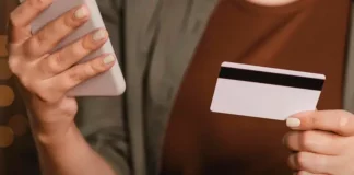 Foto mostra o tronco e as mãos de uma mulher segurando um cartão de crédito na mão esquerda e um smartphone na mão direita. A imagem simboliza o uso de tecnologia para pagamentos digitais, com foco nos objetos e nas mãos, sem mostrar o rosto da pessoa.