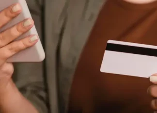 Foto mostra o tronco e as mãos de uma mulher segurando um cartão de crédito na mão esquerda e um smartphone na mão direita. A imagem simboliza o uso de tecnologia para pagamentos digitais, com foco nos objetos e nas mãos, sem mostrar o rosto da pessoa.