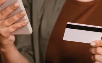 Foto mostra o tronco e as mãos de uma mulher segurando um cartão de crédito na mão esquerda e um smartphone na mão direita. A imagem simboliza o uso de tecnologia para pagamentos digitais, com foco nos objetos e nas mãos, sem mostrar o rosto da pessoa.