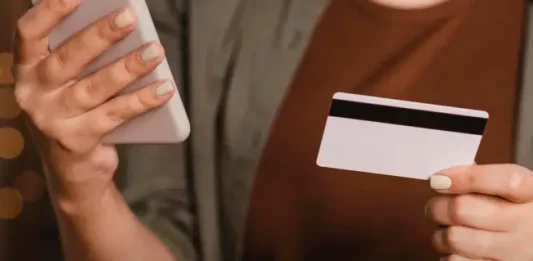 Foto mostra o tronco e as mãos de uma mulher segurando um cartão de crédito na mão esquerda e um smartphone na mão direita. A imagem simboliza o uso de tecnologia para pagamentos digitais, com foco nos objetos e nas mãos, sem mostrar o rosto da pessoa.