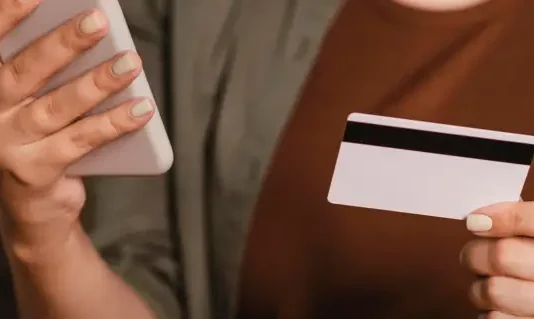 Foto mostra o tronco e as mãos de uma mulher segurando um cartão de crédito na mão esquerda e um smartphone na mão direita. A imagem simboliza o uso de tecnologia para pagamentos digitais, com foco nos objetos e nas mãos, sem mostrar o rosto da pessoa.