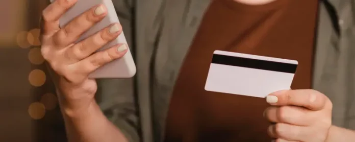 Foto mostra o tronco e as mãos de uma mulher segurando um cartão de crédito na mão esquerda e um smartphone na mão direita. A imagem simboliza o uso de tecnologia para pagamentos digitais, com foco nos objetos e nas mãos, sem mostrar o rosto da pessoa.