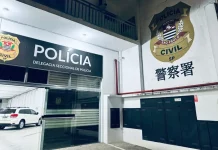 Fachada interna de uma delegacia de polícia. A imagem mostra uma porta de vidro aberta. Acima da porta, está a logo da Polícia Civil e a inscrição "Delegacia Seccional de Polícia". Do lado de fora, no pátio, há um carro branco estacionado.