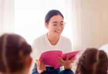 Uma professora está sentada em um puff, sorrindo enquanto segura e lê um livro de capa rosa. Ela usa camiseta branca, calça jeans azul e óculos. À sua frente, duas meninas com camisetas rosa estão sentadas, de costas para a câmera, ouvindo atentamente a leitura. A cena transmite um momento de aprendizado e acolhimento.