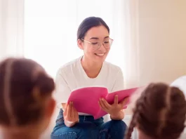 Uma professora está sentada em um puff, sorrindo enquanto segura e lê um livro de capa rosa. Ela usa camiseta branca, calça jeans azul e óculos. À sua frente, duas meninas com camisetas rosa estão sentadas, de costas para a câmera, ouvindo atentamente a leitura. A cena transmite um momento de aprendizado e acolhimento.