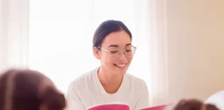 Uma professora está sentada em um puff, sorrindo enquanto segura e lê um livro de capa rosa. Ela usa camiseta branca, calça jeans azul e óculos. À sua frente, duas meninas com camisetas rosa estão sentadas, de costas para a câmera, ouvindo atentamente a leitura. A cena transmite um momento de aprendizado e acolhimento.