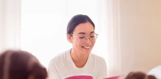 Uma professora está sentada em um puff, sorrindo enquanto segura e lê um livro de capa rosa. Ela usa camiseta branca, calça jeans azul e óculos. À sua frente, duas meninas com camisetas rosa estão sentadas, de costas para a câmera, ouvindo atentamente a leitura. A cena transmite um momento de aprendizado e acolhimento.