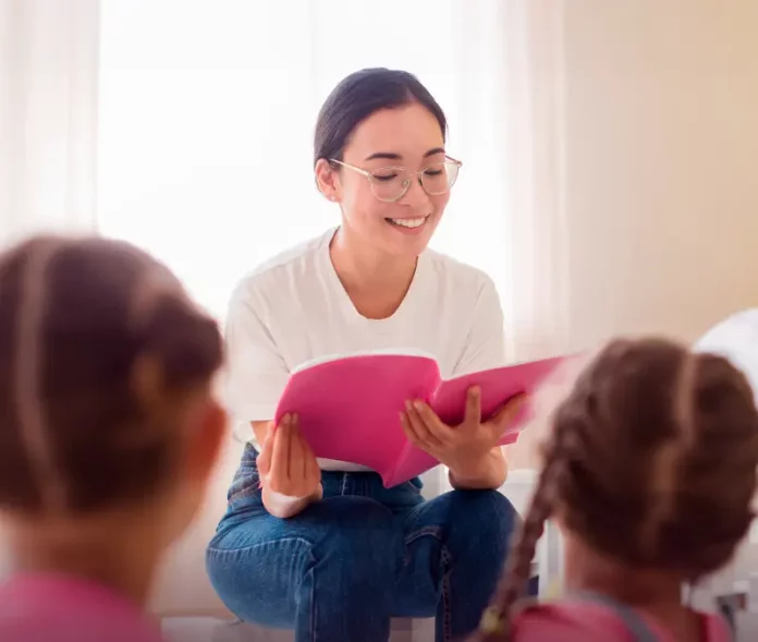 Uma professora está sentada em um puff, sorrindo enquanto segura e lê um livro de capa rosa. Ela usa camiseta branca, calça jeans azul e óculos. À sua frente, duas meninas com camisetas rosa estão sentadas, de costas para a câmera, ouvindo atentamente a leitura. A cena transmite um momento de aprendizado e acolhimento.