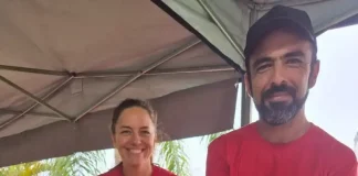 Casal de agricultores usando camisetas vermelhas atende clientes atrás do balcão da feira orgânica em São José. Na frente, há uma variedade de produtos frescos e naturais, como alface, repolho roxo, repolho verde, cenoura, batata-doce, rabanete e banana, destacando a diversidade da agricultura familiar local.