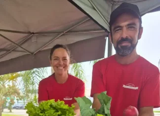 Casal de agricultores usando camisetas vermelhas atende clientes atrás do balcão da feira orgânica em São José. Na frente, há uma variedade de produtos frescos e naturais, como alface, repolho roxo, repolho verde, cenoura, batata-doce, rabanete e banana, destacando a diversidade da agricultura familiar local.